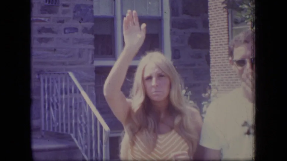 A closer home-movie frame shows the woman holding her hand raised, with the man partly visible near the edge and building steps behind her.