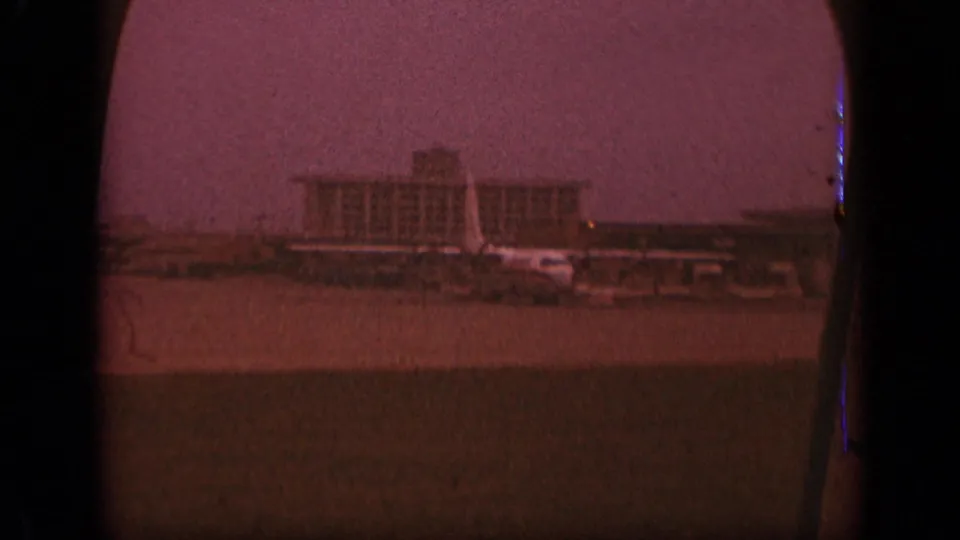1970 New Jersey airport terminal and tarmac viewed through an airplane window on grainy 8mm film.