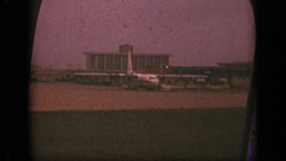 Passenger view of a New Jersey airport apron with a terminal building and aircraft tail visible in 1970 8mm footage.