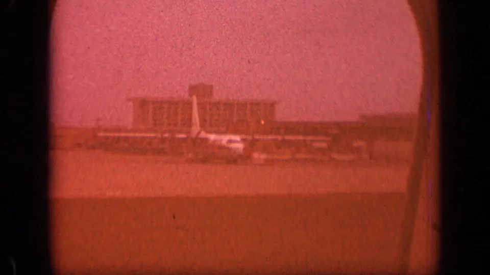 Red-shifted 8mm passenger-window view of a New Jersey airport terminal, apron, and aircraft tail in 1970.