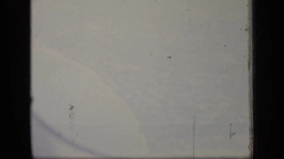 Hazy 1970 8mm airplane-window view with a pale sky, dark window edge, dust spots, and vertical scratch marks.