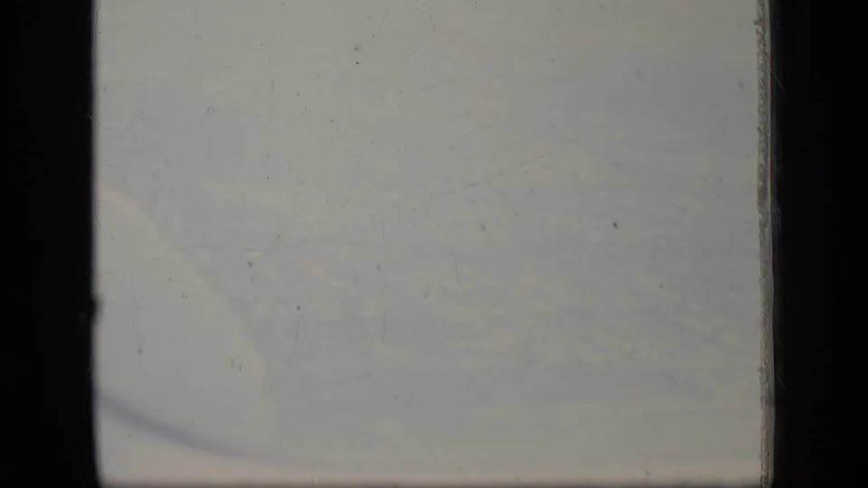 Final 8mm frame of the New Jersey flight view, showing hazy sky, faint cloud forms, window edges, grain, and dust.