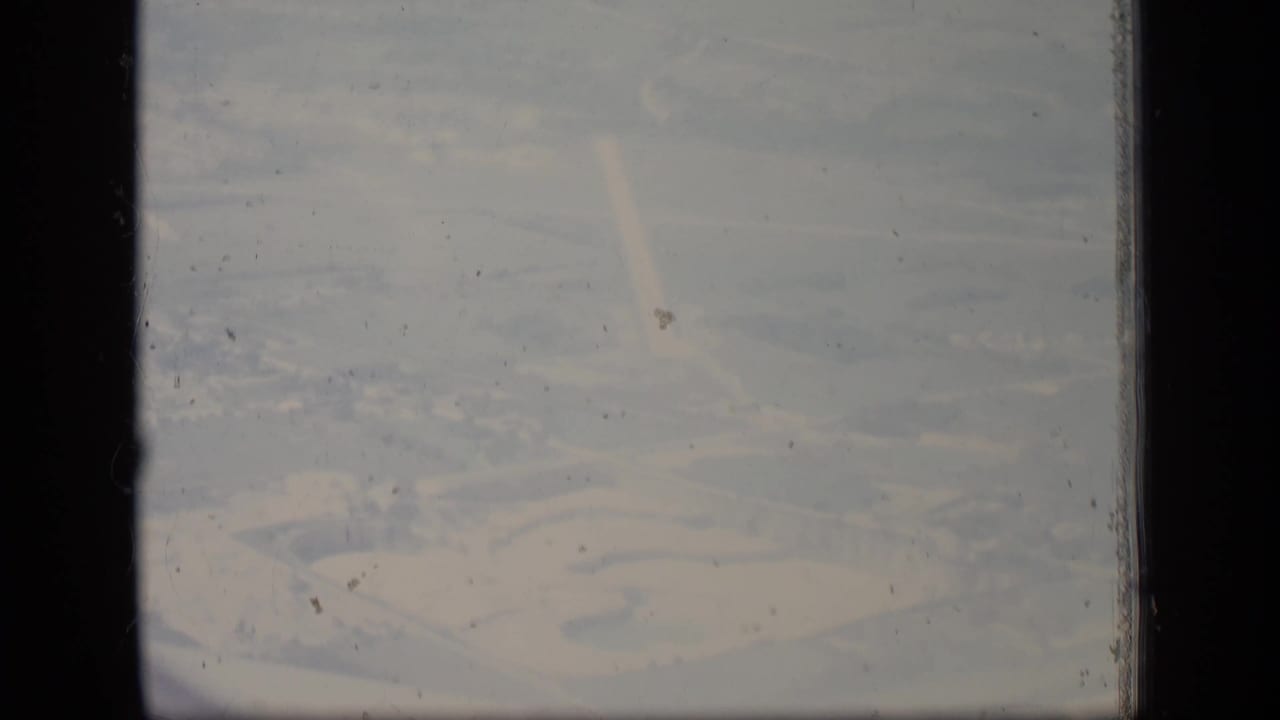 Hazy aerial view of terrain and a large facility area seen through 8mm film gate edges.