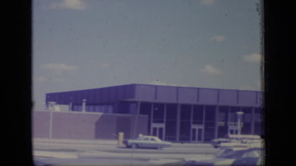 Ground-level 1970 New Jersey frame showing the facility entrance, glass bays, parked cars, blue sky, and faded 8mm color.