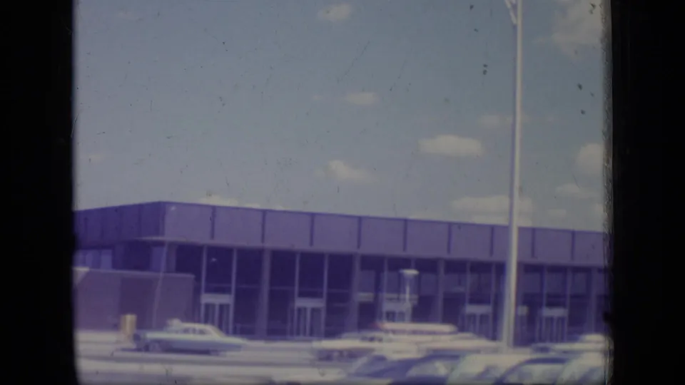 1970 New Jersey 8mm frame showing a low modernist commercial or residential building along a street with classic cars nearby.