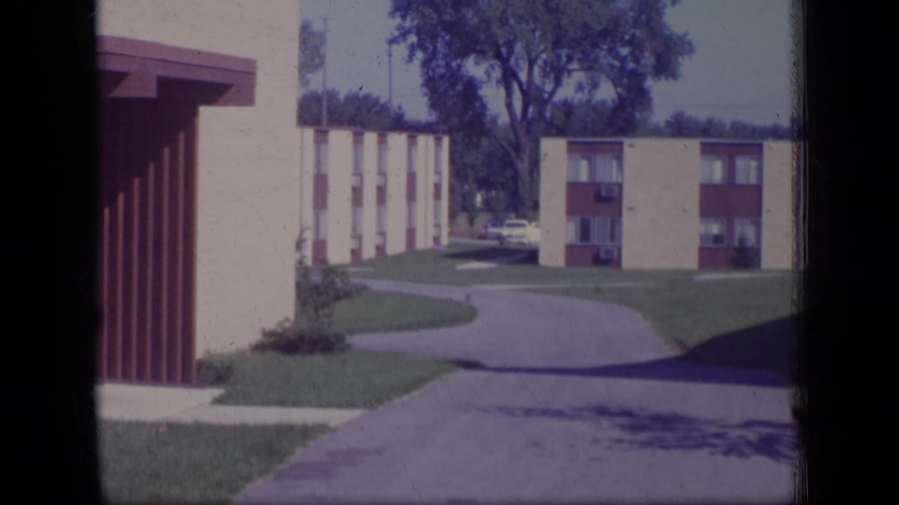 1970 New Jersey home-movie frame of low-rise apartment buildings, a curved driveway, lawns, and a mature tree.