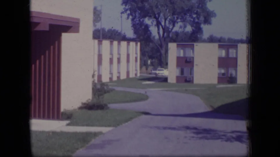 1970 New Jersey home-movie frame of low-rise apartment buildings, a curved driveway, lawns, and a mature tree.