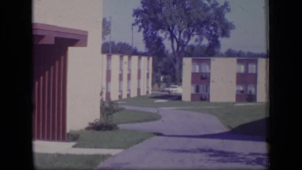 1970 New Jersey 8mm frame showing cream and brown apartment blocks beside a driveway and maintained lawns.