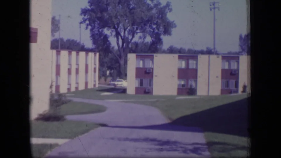 1970 New Jersey home-movie frame of an apartment-complex driveway, low-rise buildings, lawns, trees, and parked cars.