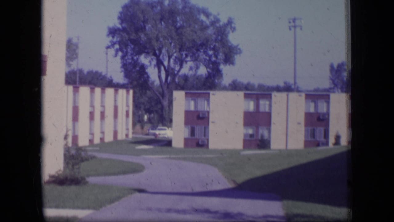 1970 New Jersey home-movie frame of low-rise apartment buildings, lawn, curving driveway, and a large tree behind the complex.