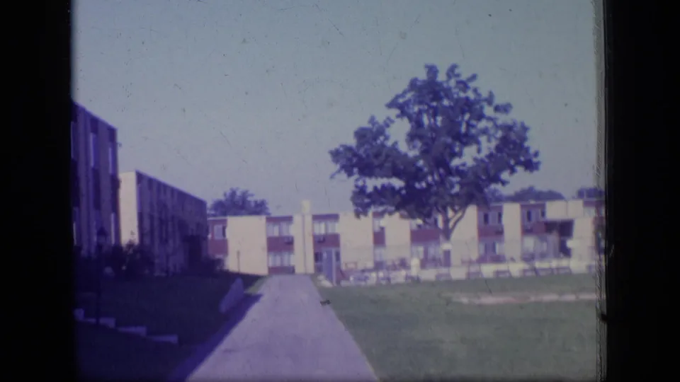 1970 New Jersey 8mm frame looking down a paved drive toward apartment buildings, lawns, parked cars, and a central leafy tree.