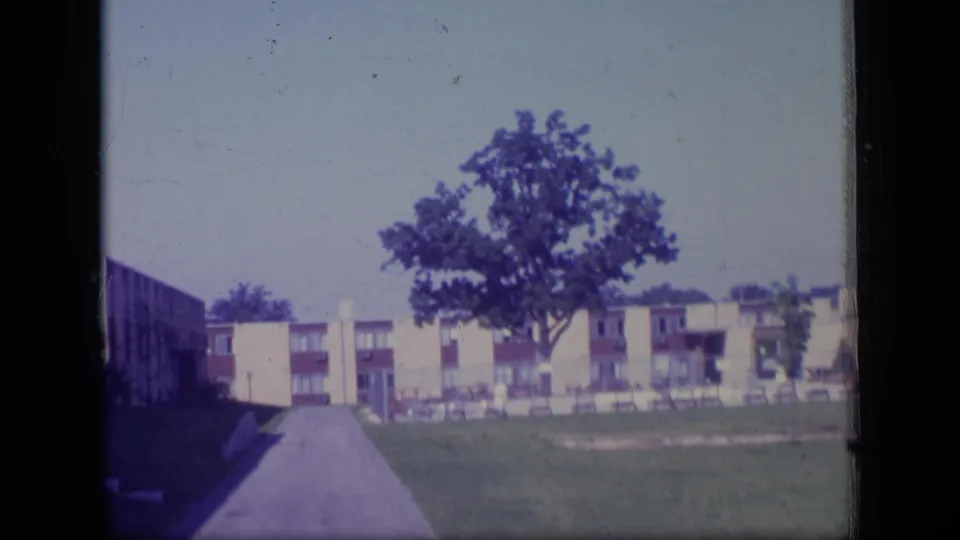 1970 New Jersey home-movie view of a quiet apartment complex with a paved drive, central tree, lawn, and low-rise buildings.