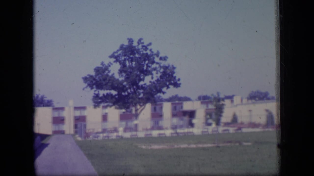 Static 8mm view of a New Jersey residential lawn with a broad tree, walkway, and low-rise apartment facade, circa 1970.