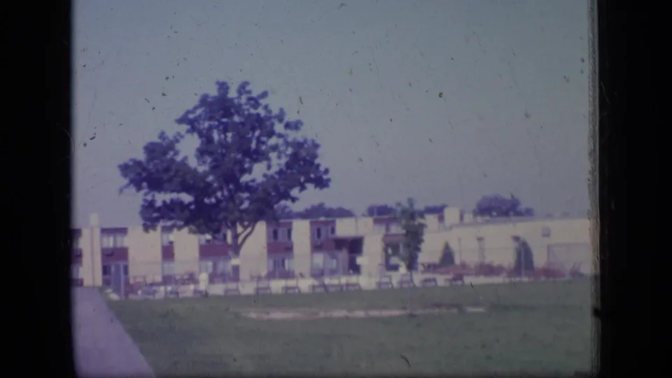 1970 home-movie frame from New Jersey showing a leafy tree, open grass, and a long low-rise residential structure.
