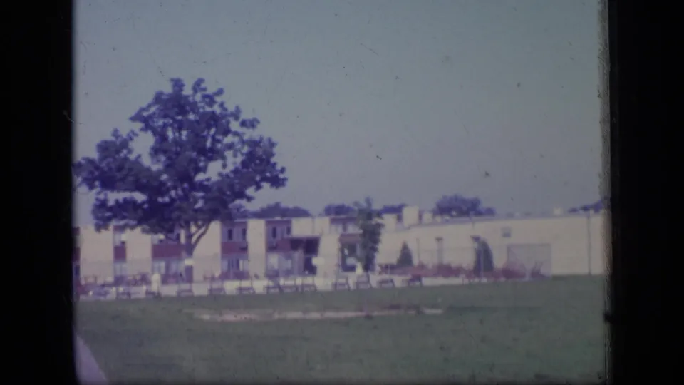 Soft 8mm frame of a New Jersey suburban exterior in 1970, with lawn, tree, and low-rise residential buildings under a pale sky.