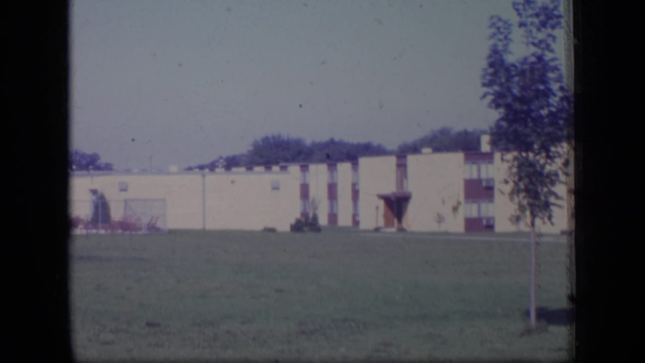 1970 New Jersey home-movie frame with a long cream building and red details seen beyond an open lawn.