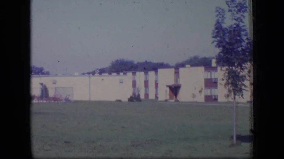 1970 New Jersey home-movie frame with a long cream building and red details seen beyond an open lawn.