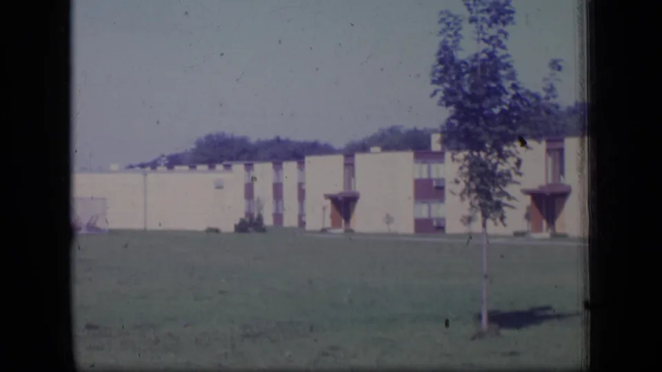 1970 New Jersey 8mm frame showing a young planted tree before a cream low-rise building on a lawn.