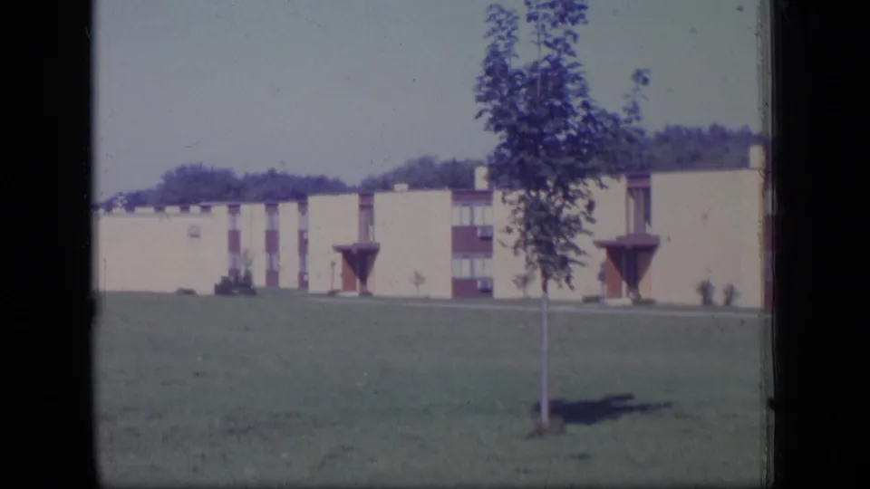 Final 1970 New Jersey frame with a foreground tree, grassy field, and cream low-rise building with red accents.