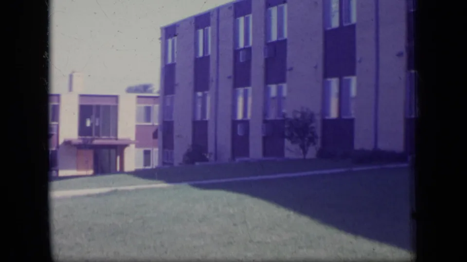 1970 New Jersey home-movie frame showing a low-rise apartment facade, lawn, walkway, and shaded grass.
