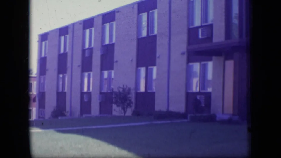1970 New Jersey 8mm view of an apartment building corner, windows, small shrub, and open lawn.