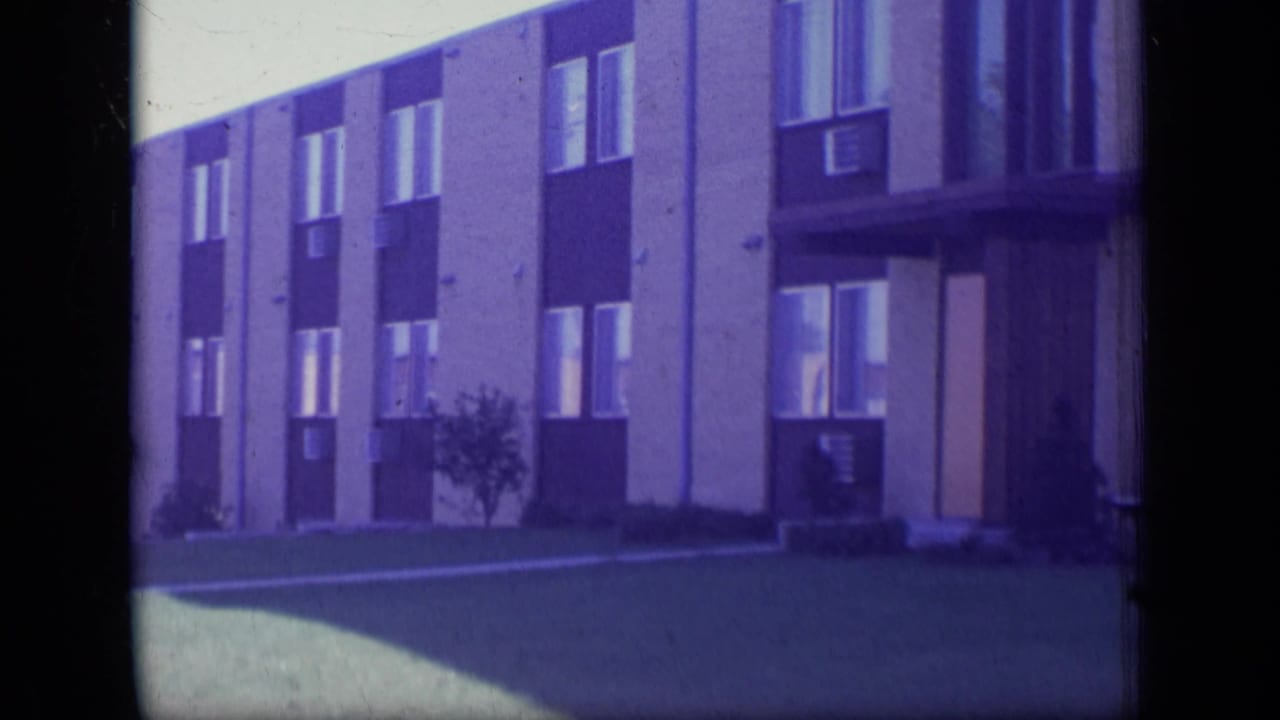 1970 New Jersey 8mm view of a beige and dark-panel apartment building with windows, air conditioners, lawn, and a small tree.
