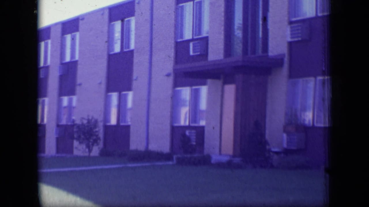1970 New Jersey home-movie frame showing the building entrance, rows of windows, air conditioners, lawn, and shrubs.