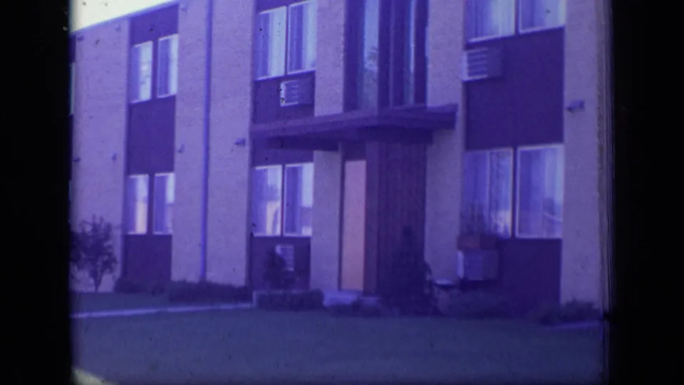 Closer 1970 New Jersey 8mm frame of the apartment entrance with dark facade panels, windows, air conditioners, and lawn.