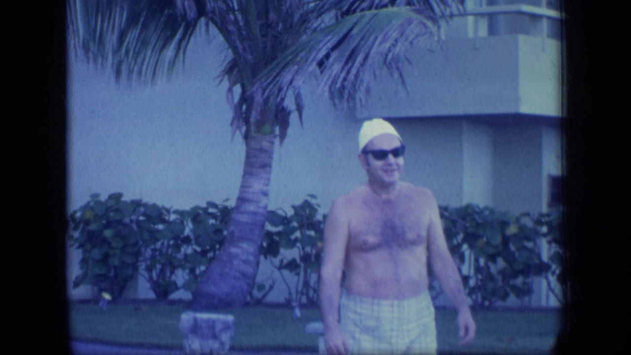 In 1970 Puerto Rico home-movie footage, a shirtless man in a yellow swim cap and sunglasses stands outdoors by palms and a modern building.