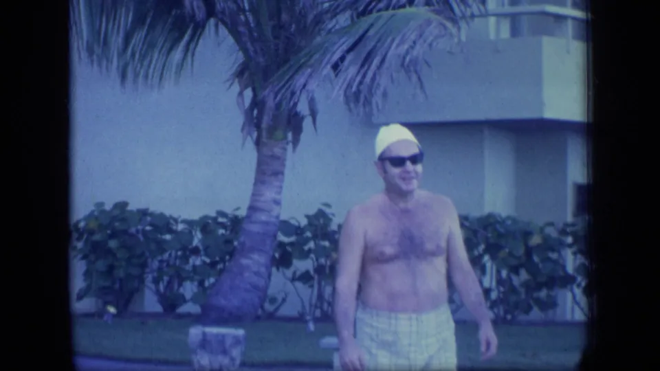 In 1970 Puerto Rico home-movie footage, a shirtless man in a yellow swim cap and sunglasses stands outdoors by palms and a modern building.