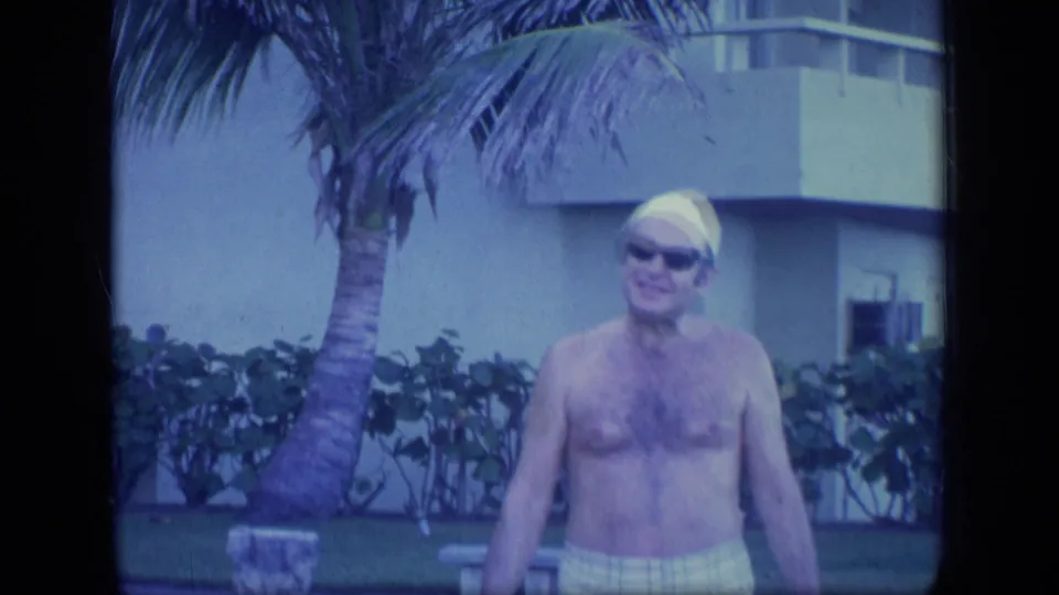 The man walks toward the camera in 1970 Puerto Rico, wearing sunglasses, a yellow swim cap and plaid trunks in a tropical resort setting.