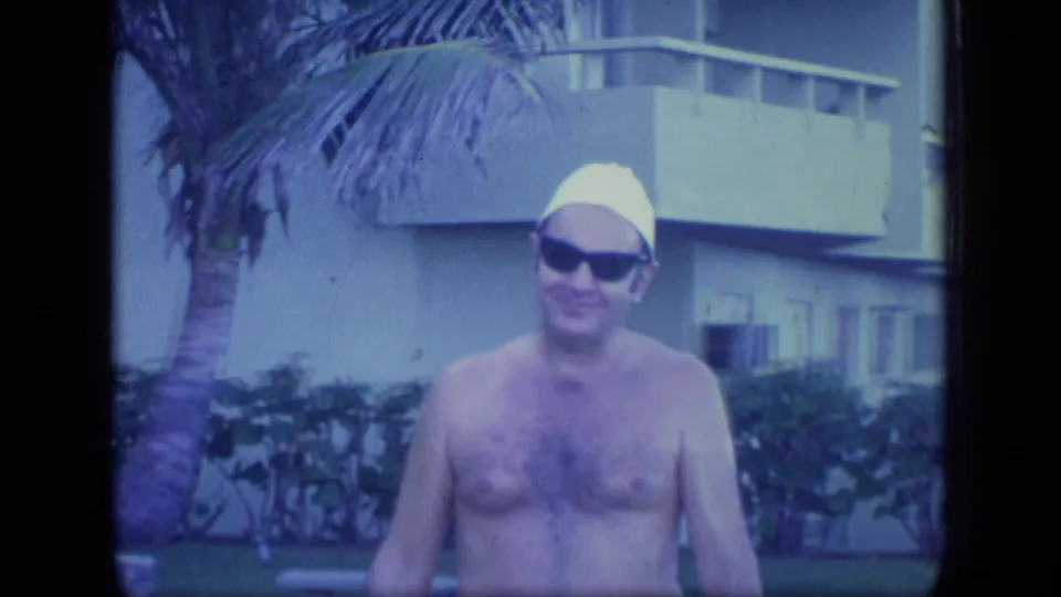 Closer 8mm frame of the shirtless man in sunglasses and a yellow swim cap, with palm landscaping and a resort-style building behind him.