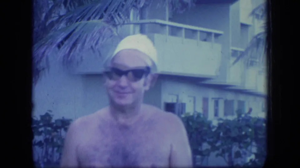 Close view of the smiling man in a yellow swim cap and dark sunglasses, filmed outdoors in Puerto Rico with palms and a pale building visible.