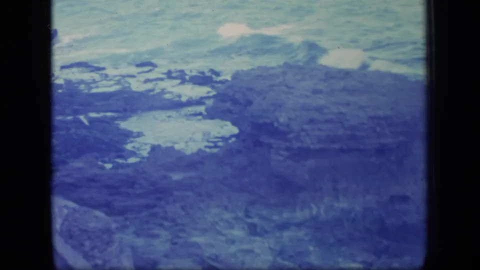 Final 1970 Puerto Rico coastal frame with waves, a broad rock outcrop, shallow pools, and foreground grasses.