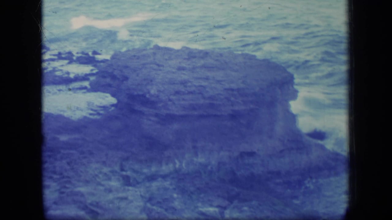 Rocky shoreline with blue water and white surf seen through an 8mm film frame.