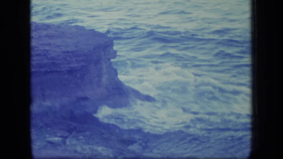 1970 Puerto Rico 8mm frame of waves pressing against a rugged rock face at the shoreline.