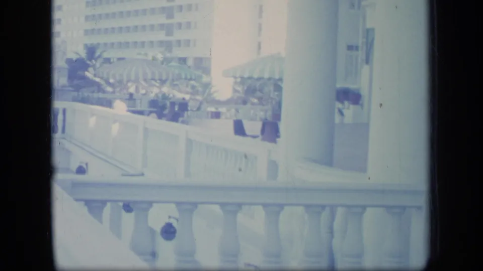1970 Puerto Rico balcony view toward a resort terrace with striped umbrellas, white railings, plants, and a tall building beyond.