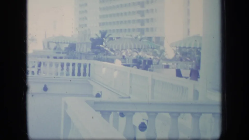 Soft 8mm frame of a Puerto Rico resort balcony and terrace, with striped umbrellas, white railings, plants, and a large building in view.