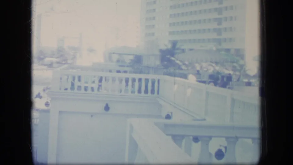 1970 Puerto Rico home-movie frame looking across a bright resort balcony area with white railings, striped umbrellas, plants, and distant people.