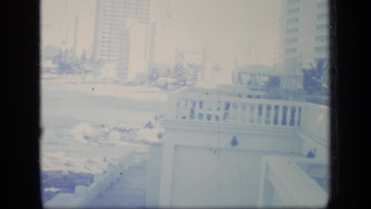 Rocky surf and balcony edge with distant beachfront buildings and palm trees.