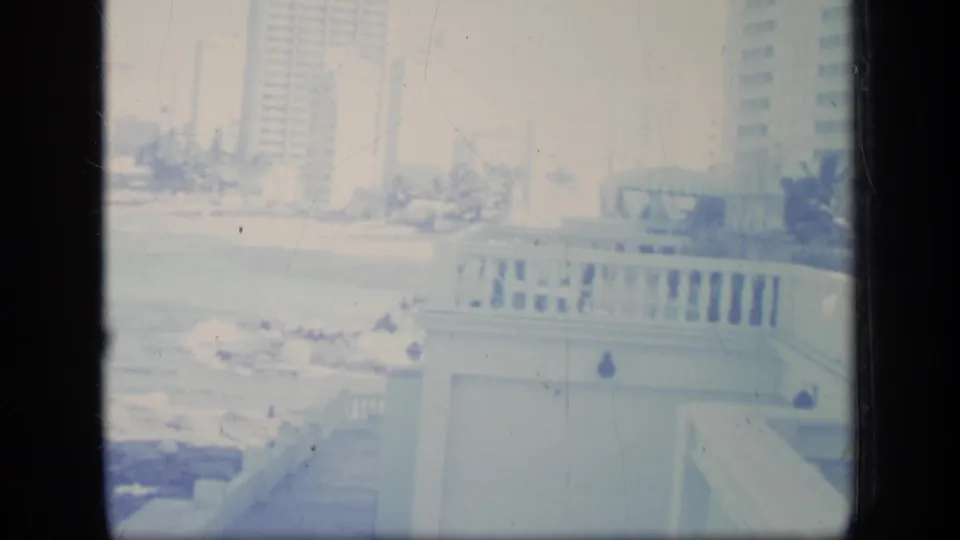 1970 Puerto Rico home-movie frame with surf breaking on rocks below a terrace railing and beachfront towers in the distance.