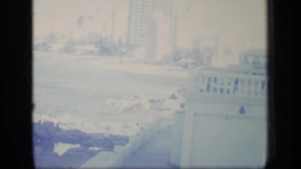 1970 Puerto Rico coastal pan frame showing pale ocean water, dark rocks, a balcony railing, palms, and tall resort-like buildings.