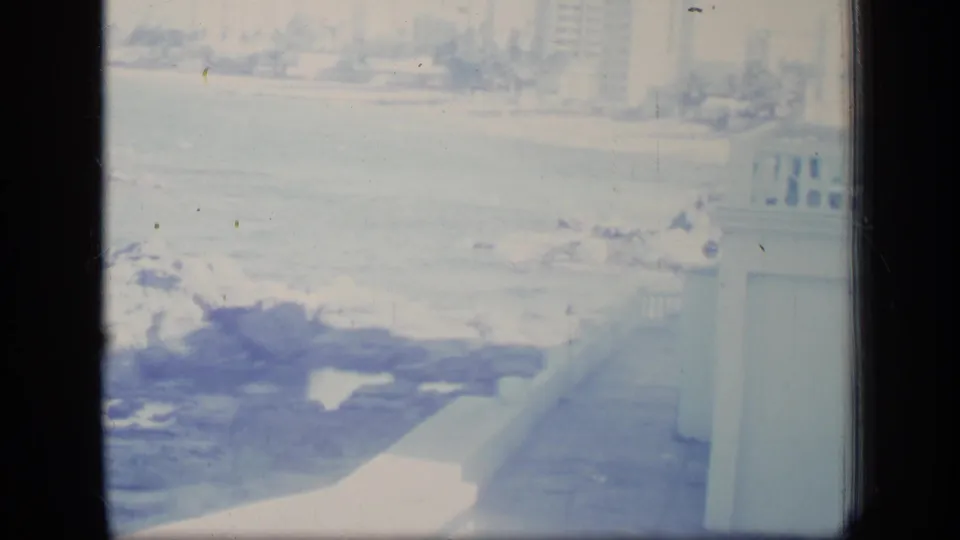 1970 Puerto Rico 8mm frame with foamy surf over rocks, a balcony rail at right, and distant beach activity in bright exposure.