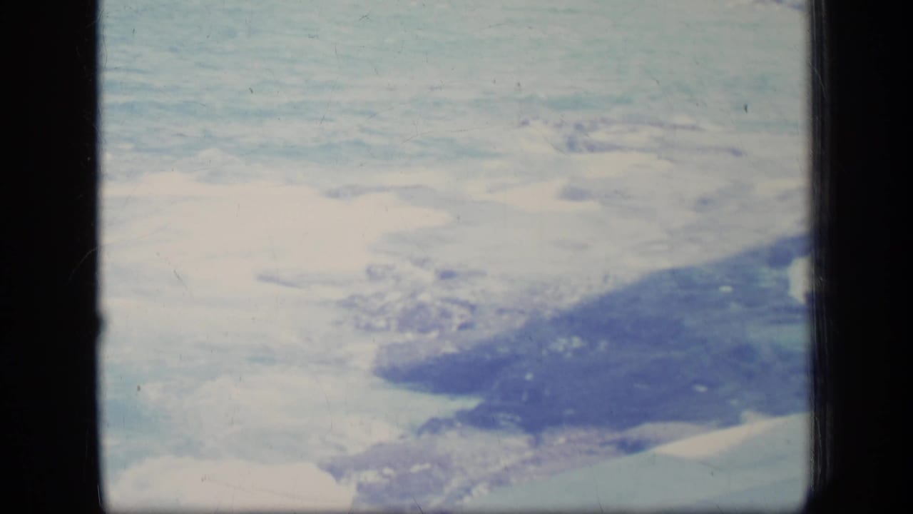1970 8mm airplane-window view of snow-covered terrain, dark shoreline forms, and pale water or ice below.