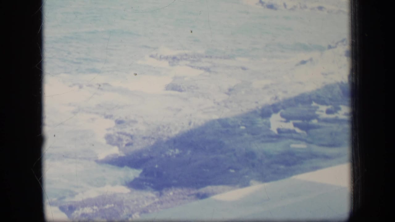 1970 8mm aerial frame showing snow-bright ground, darker coastal landforms, and soft blue-green film color.