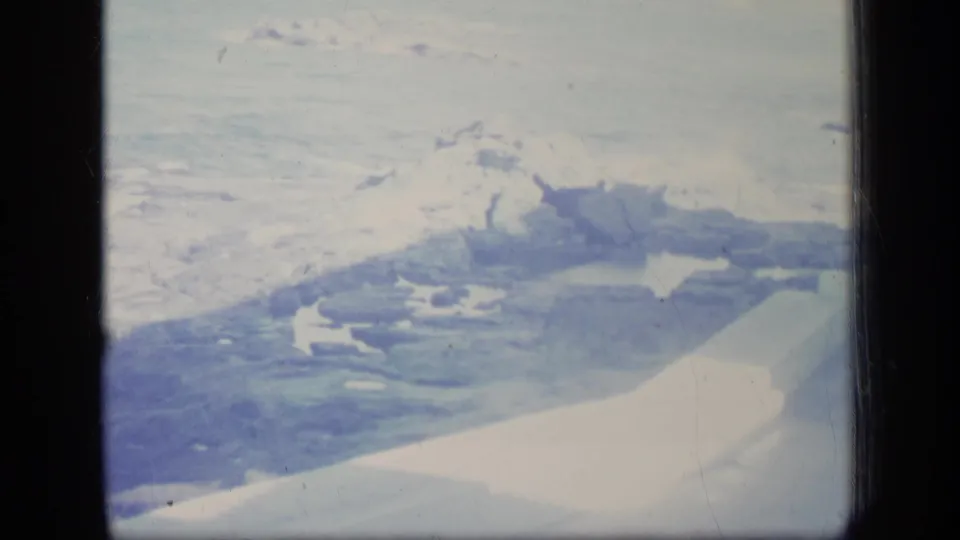1970 8mm frame of an airplane wing crossing a snowy shoreline or mountain landscape seen from above.
