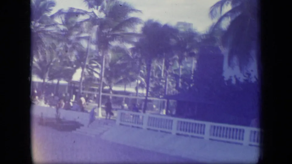 Puerto Rico beach scene from a 1970 8mm home movie showing palms, a white railing, and distant people near resort-style buildings.