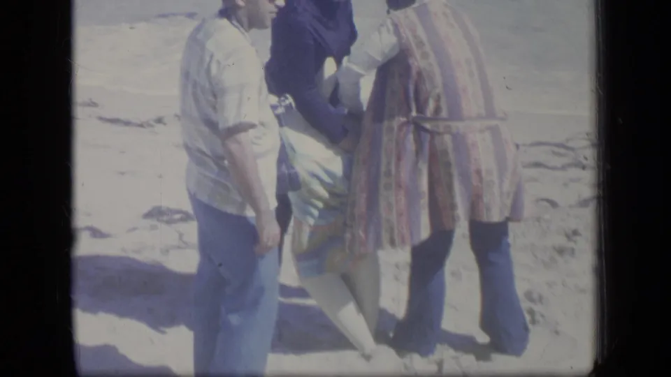 A 1970 Puerto Rico beach frame shows adults in casual clothing gathered closely beside a person low on the sand.