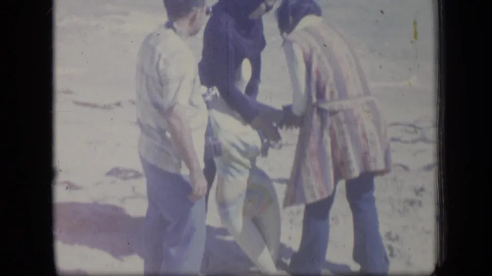 The 1970 Puerto Rico home-movie frame shows adults standing and leaning near someone low on a bright sandy beach.