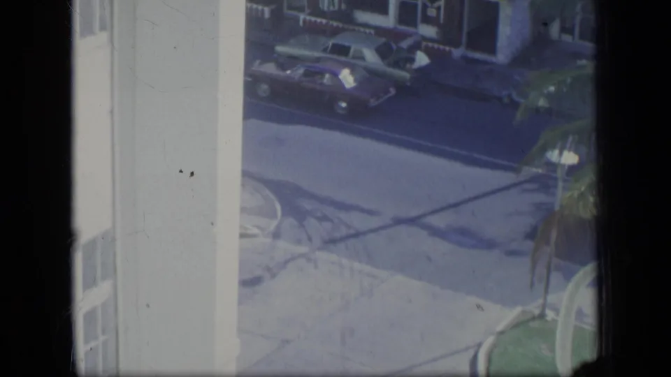 1970 Puerto Rico street scene seen from a high window, with vintage cars near storefronts and palm fronds.